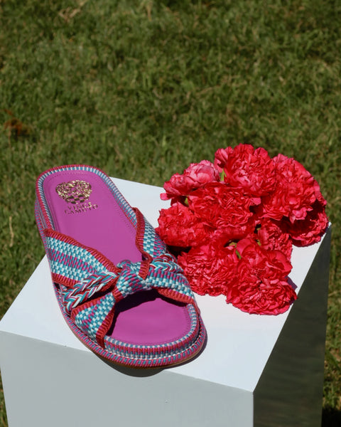 The Vince Camuto Gryfynn Slide, featuring a colorful woven upper, cushioned sole, and purple footbed, is displayed on a white pedestal with red carnations and lush green grass in the background for a fresh summer vibe.