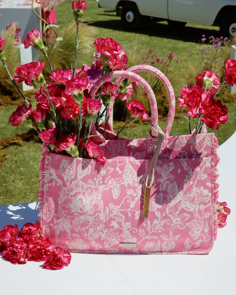 A pink, floral-patterned Zhury Tote Bag by Vince Camuto filled with bright pink carnations sits on a white surface outdoors, with grassy lawn and a white vehicle softly blurred in the background.