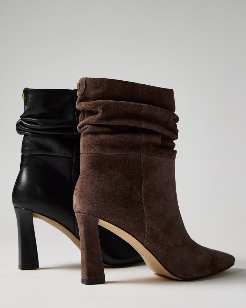 Two Grenna Slouchy Bootie high-heeled ankle boots by Vince Camuto are displayed; one is crafted from black leather, and the other features brown suede. Both styles have a slouchy silhouette with pointed toes, set against a plain light background.