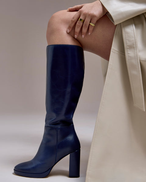 A person wearing a cream trench coat and Vince Camuto’s Lucina Knee High Boots with a block heel sits with one leg crossed, showing gold rings on their fingers.