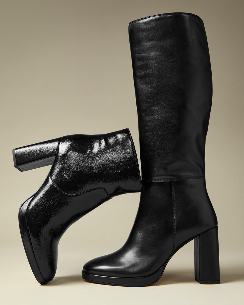 The Vince Camuto Lucina Wide Calf Knee High Boot is pictured in glossy black leather, featuring chunky block heels. One boot stands upright while the other is balanced horizontally on top, all set against a neutral backdrop. These elegant knee-high boots are designed to make a statement.