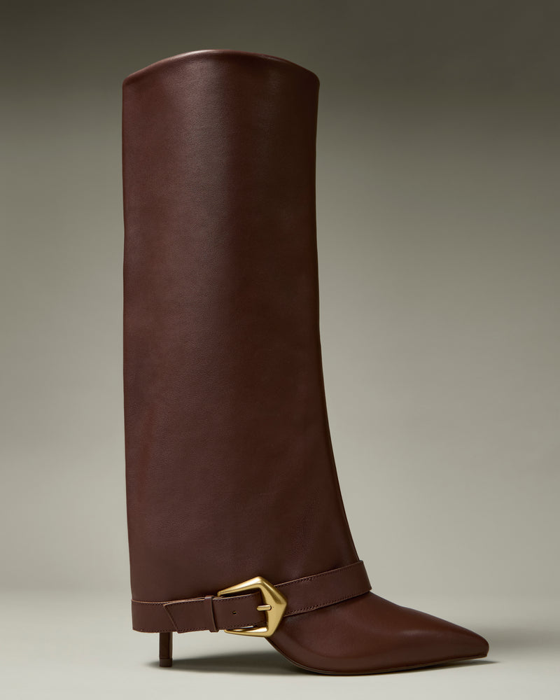 The Margee Cuff Knee High Boot by Vince Camuto is a tall, brown leather boot featuring a pointed toe, low heel, and a gold buckle strap detail at the ankle, displayed against a neutral background.