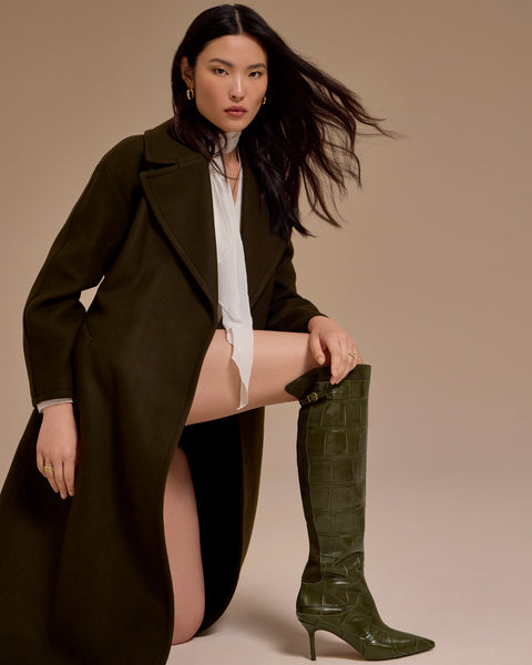 A woman poses on one knee in a dark green coat and the Vince Camuto Meleis Over the Knee Boots. She wears a white shirt and gold jewelry, with her long dark hair flowing to the side against a beige background.