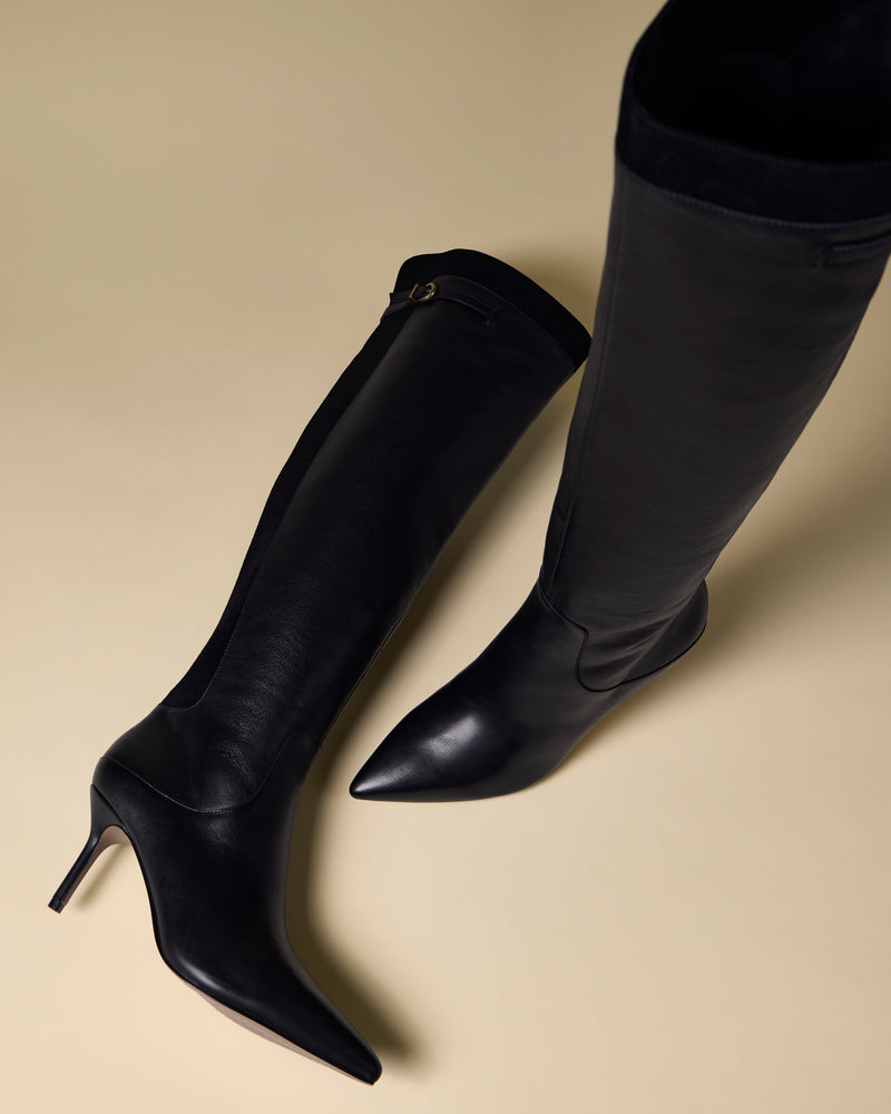 Two Meleis Over the Knee Boots by Vince Camuto are displayed on a beige background, one standing upright and the other lying down, highlighting their sleek black design with pointed toes and stiletto heels.