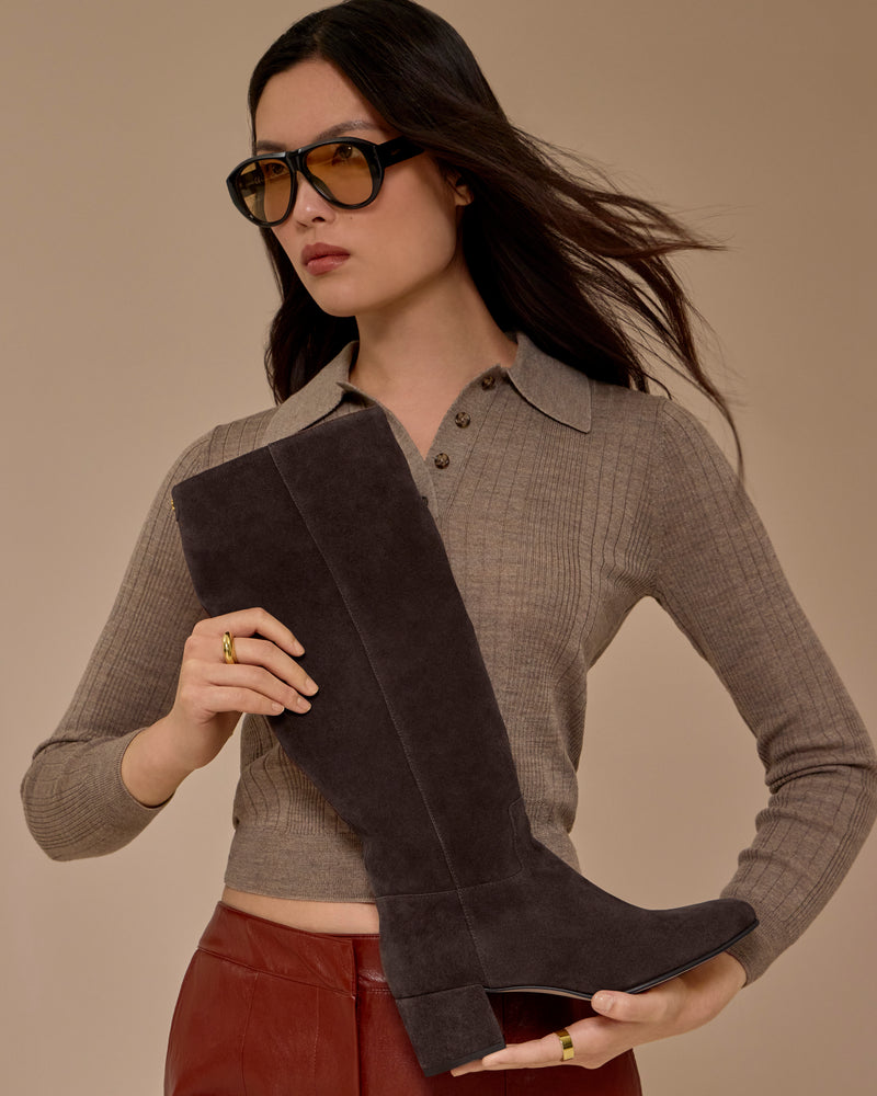 A woman in sunglasses and a taupe sweater holds the Vince Camuto Pazzla Knee High Boot in dark suede. She has long hair and wears brown pants, standing against a neutral beige background.