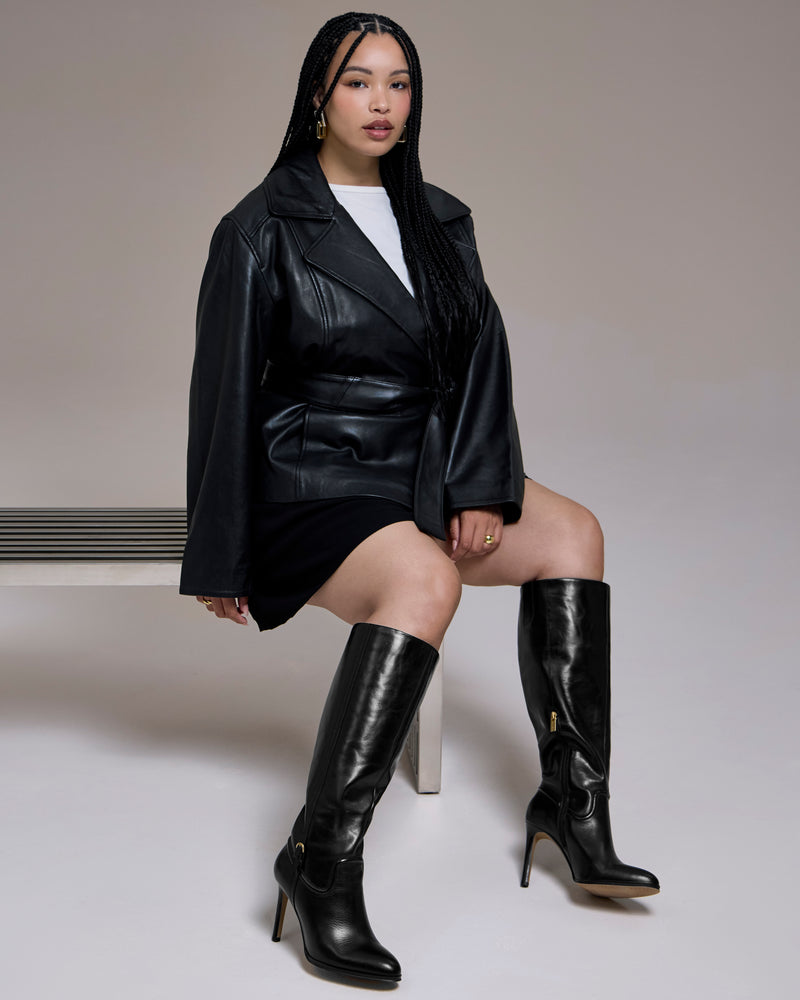 A woman with box braids sits on a bench wearing a black leather jacket, white top, black skirt, and the Vince Camuto Sareya Knee High Boot—crafted from premium materials—which creates a sleek silhouette against a plain background.