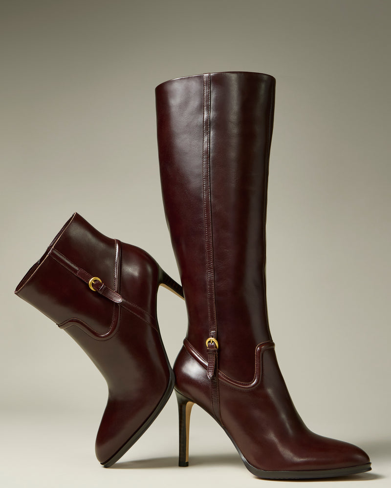 A pair of Sareya Knee High Boots by Vince Camuto, crafted from premium brown leather and adorned with gold buckle accents; one boot stands upright while the other leans gracefully against it, highlighting their sleek silhouette.