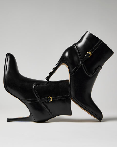 Two Vince Camuto Setily Bootie high-heeled ankle boots are balanced heel to toe against each other on a plain gray background, highlighting their sleek black leather texture and gold buckle accents.