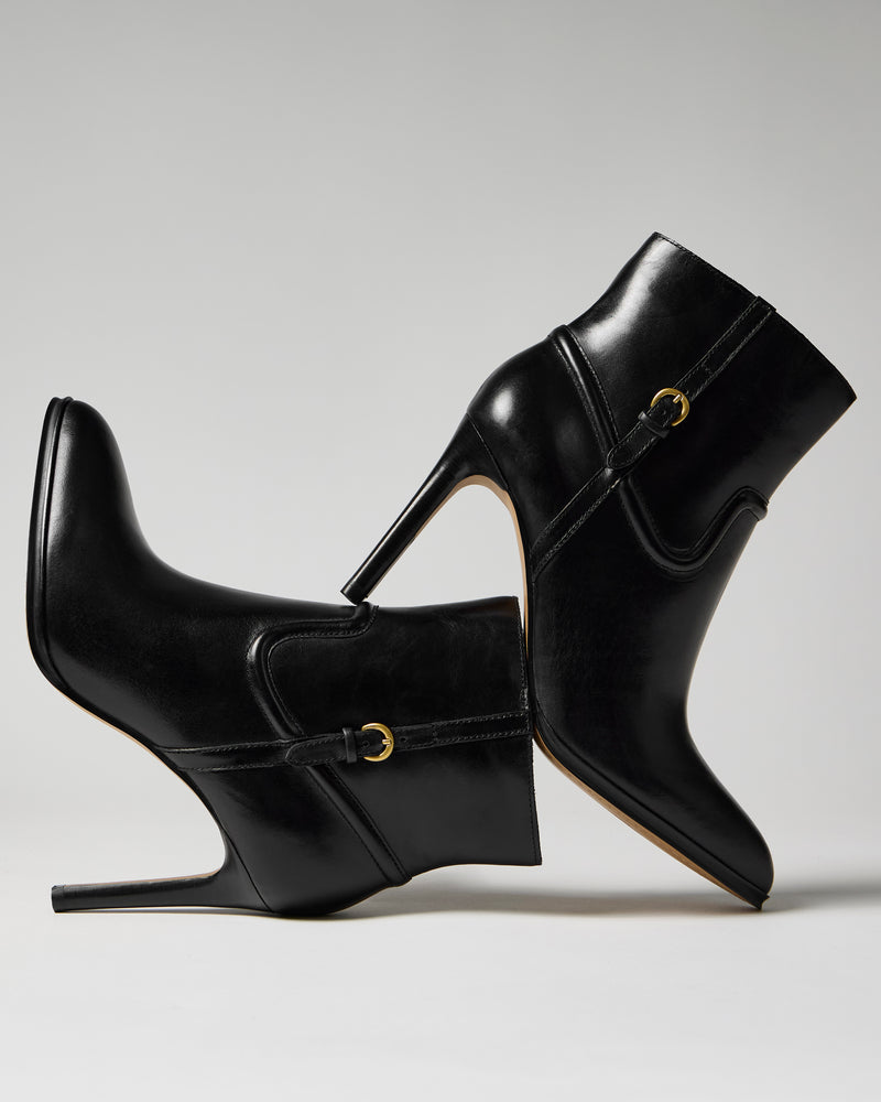 Two Vince Camuto Setily Bootie high-heeled ankle boots are balanced heel to toe against each other on a plain gray background, highlighting their sleek black leather texture and gold buckle accents.