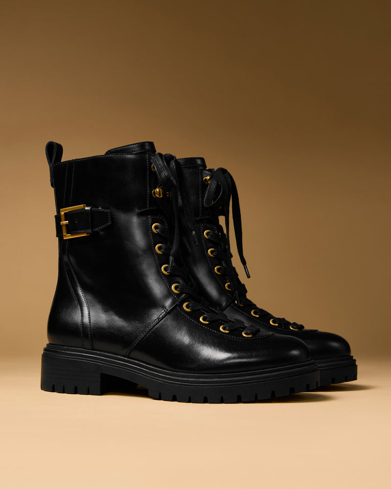 The Vince Camuto Becinda Moto Bootie, a pair of black leather lace-up combat boots featuring gold eyelets and a decorative side buckle, is displayed on a beige surface with a brown gradient background.