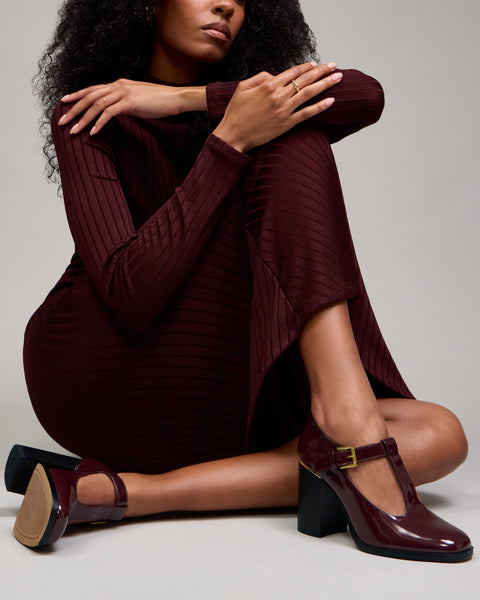 A woman is seated on the floor in a burgundy ribbed dress, legs crossed, wearing Vince Camuto’s Micha Pump—a pair of matching chunky-heeled Mary Jane shoes with gold buckles that evoke vintage charm. Her hands rest on her knee, her face partly out of frame, against a plain gray background.