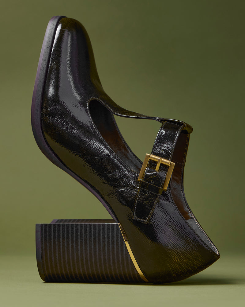 The Vince Camuto Micha Pump, a black patent leather Mary Jane shoe featuring a chunky, striped wedge heel and an elegant gold buckle, is displayed on its side against an olive green background—beautifully capturing vintage charm while embracing feminine style.