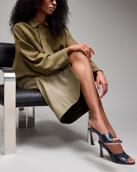 A woman sits on a modern chair, wearing an olive coat, beige skirt, and shiny Vince Camuto Paigely Mule sandals with a flared heel. Her legs are crossed, and she has curly hair. Only the lower part of her face and body are visible.