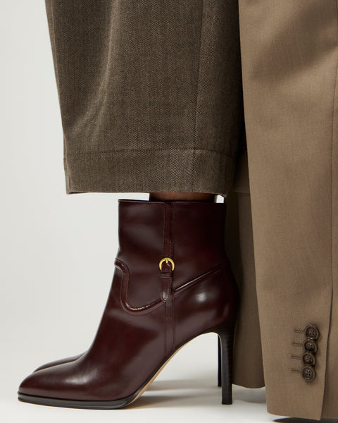 A person wearing brown trousers and a beige coat stands in Vince Camuto Setily Bootie dark brown heeled ankle boots with a gold buckle detail.