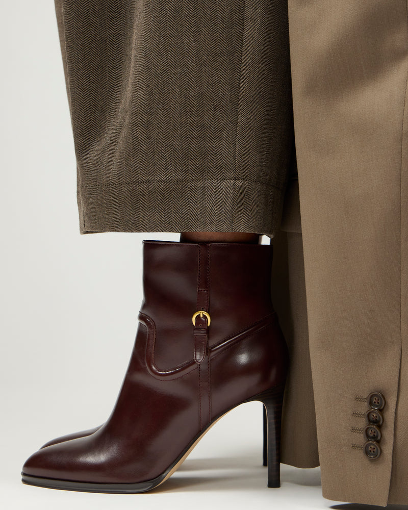 A person wearing brown trousers and a beige coat stands in Vince Camuto Setily Bootie dark brown heeled ankle boots with a gold buckle detail.