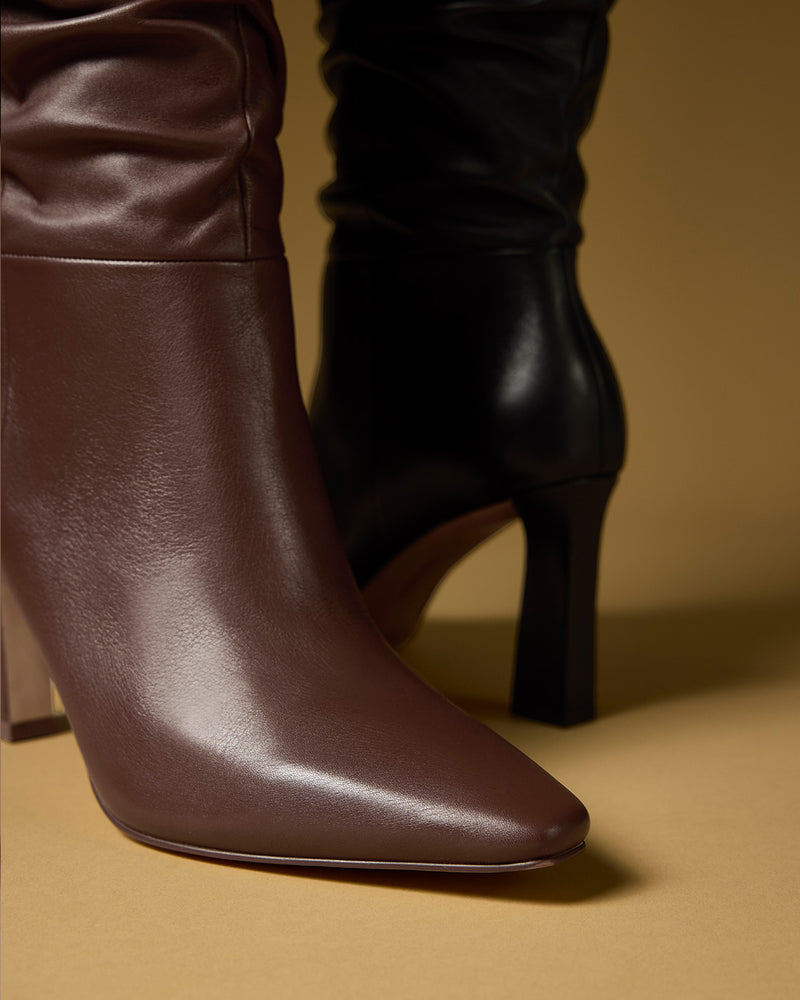 Close-up of two Vince Camuto Genisa Slouchy Knee High Boots on a tan surface, one in brown and one in black, both featuring smooth leather with a slightly pointed toe and block heel.