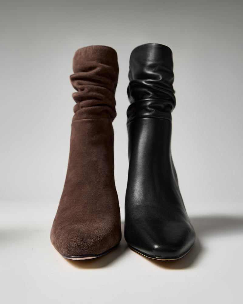 Two Vince Camuto Grenna Slouchy Booties are shown side by side; the left boot features brown suede, while the right boot features black leather. Both showcase a slouchy design with pointed toes, set against a plain gray background.