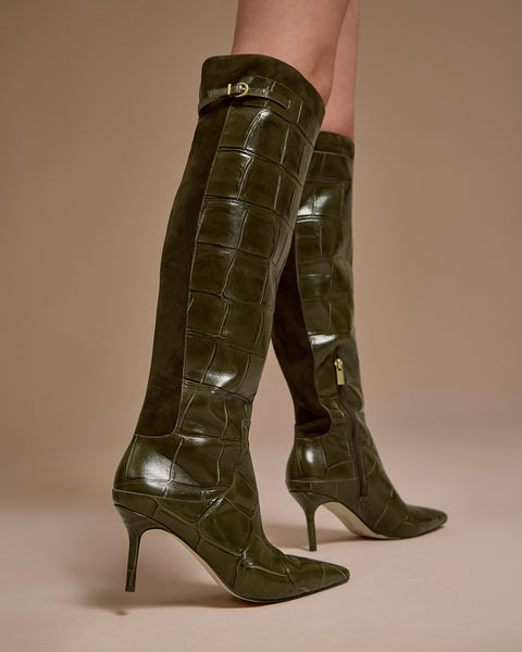 A person wearing the Vince Camuto Meleis Narrow Calf Over the Knee Boot, featuring knee-high dark green crocodile-textured leather with suede panels, gold hardware, and pointed toes, stands against a beige background.