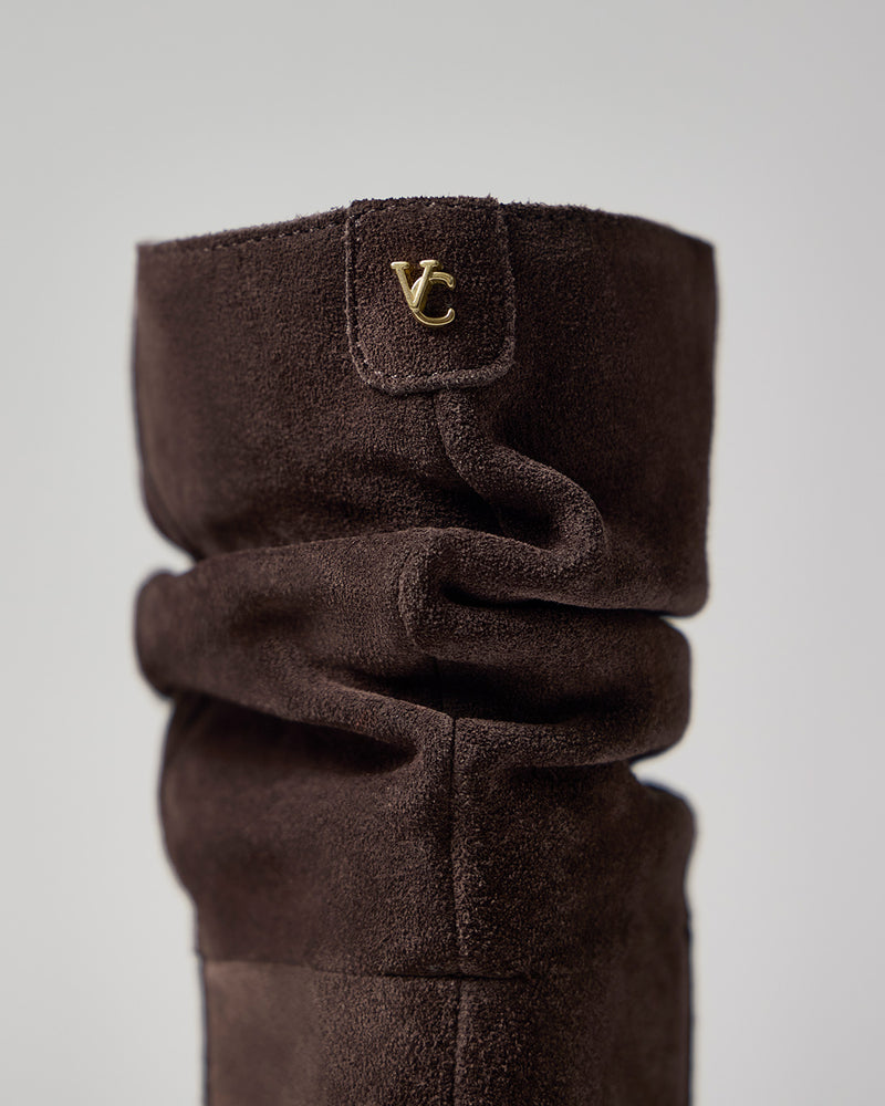 Close-up of the Vince Camuto Genisa Wide Calf Slouchy Knee High Boot in brown suede, showcasing a gold VC logo on the upper side and a slouchy, textured design set against a plain, light background.