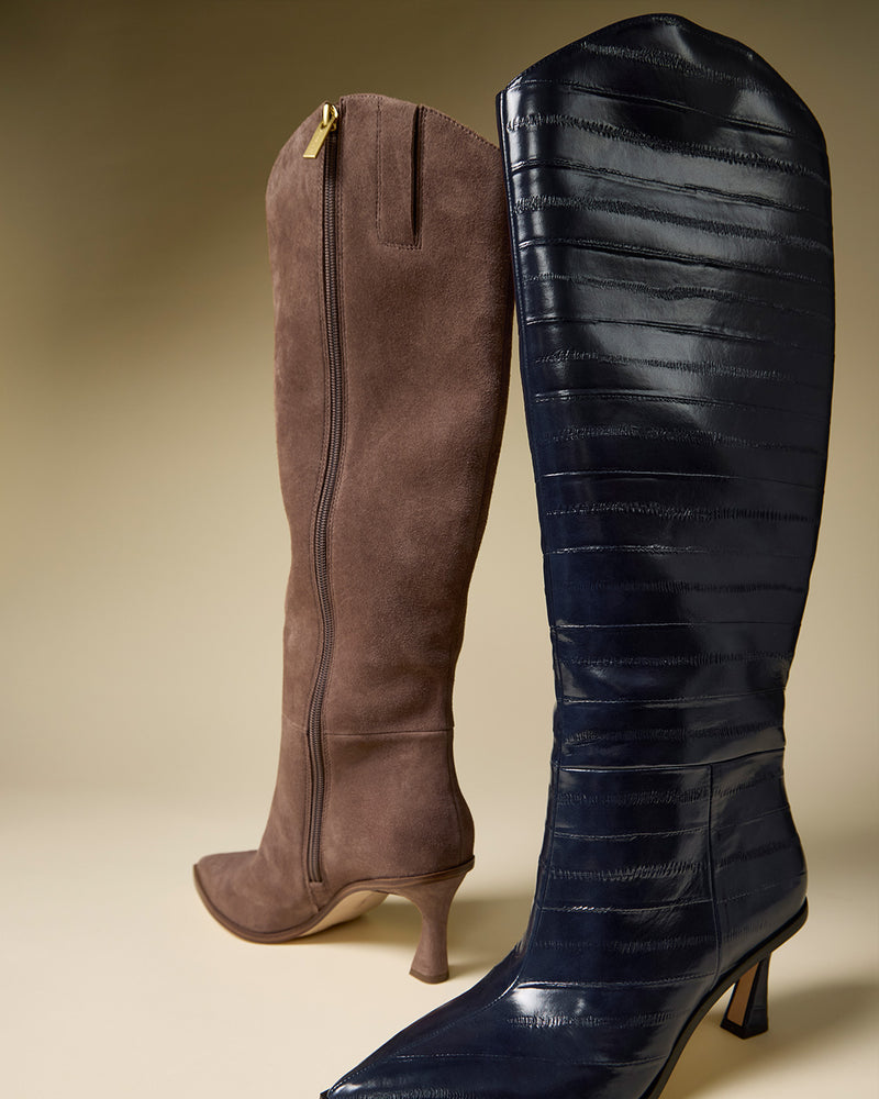 Two Vince Camuto Kenlea Narrow Calf Knee High Boots stand side by side: one in brown suede with a zipper and curved heel, the other in shiny dark blue with a subtle pattern and block heel, both upright against a neutral background.