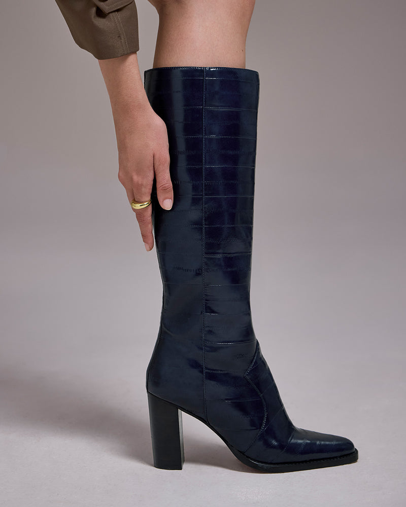 A person’s hand touches the side of a dark navy Vince Camuto Lecheya Extra Wide Calf Knee High Boot, a knee-high style with a textured finish and a block heel.