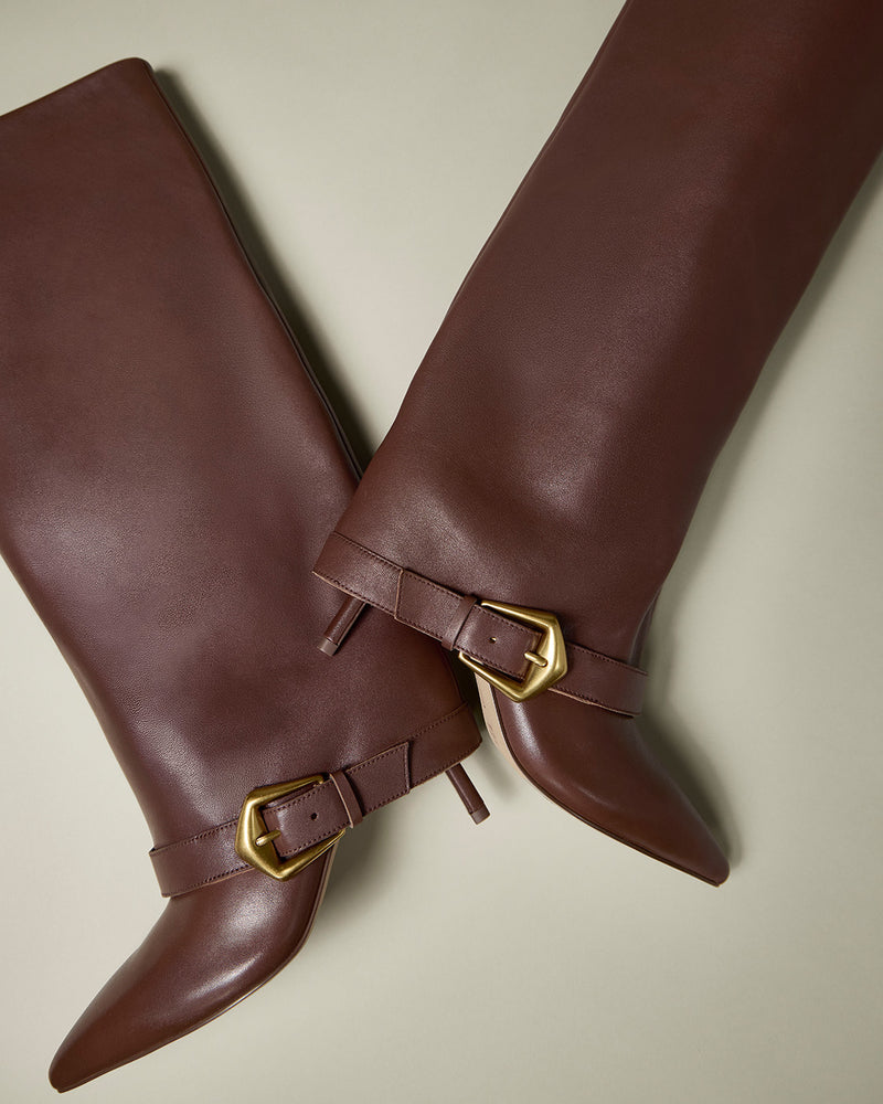 A pair of Vince Camuto Margee Wide Calf Cuff Knee High Boots in brown leather, featuring pointed toes and gold ankle buckles, positioned diagonally on a light beige background.