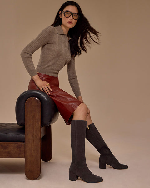 A woman with long dark hair wearing glasses, a taupe ribbed sweater, a red leather skirt, and Vince Camuto Pazzla Wide Calf Knee High Boots sits on a modern brown and black chair against a beige background.