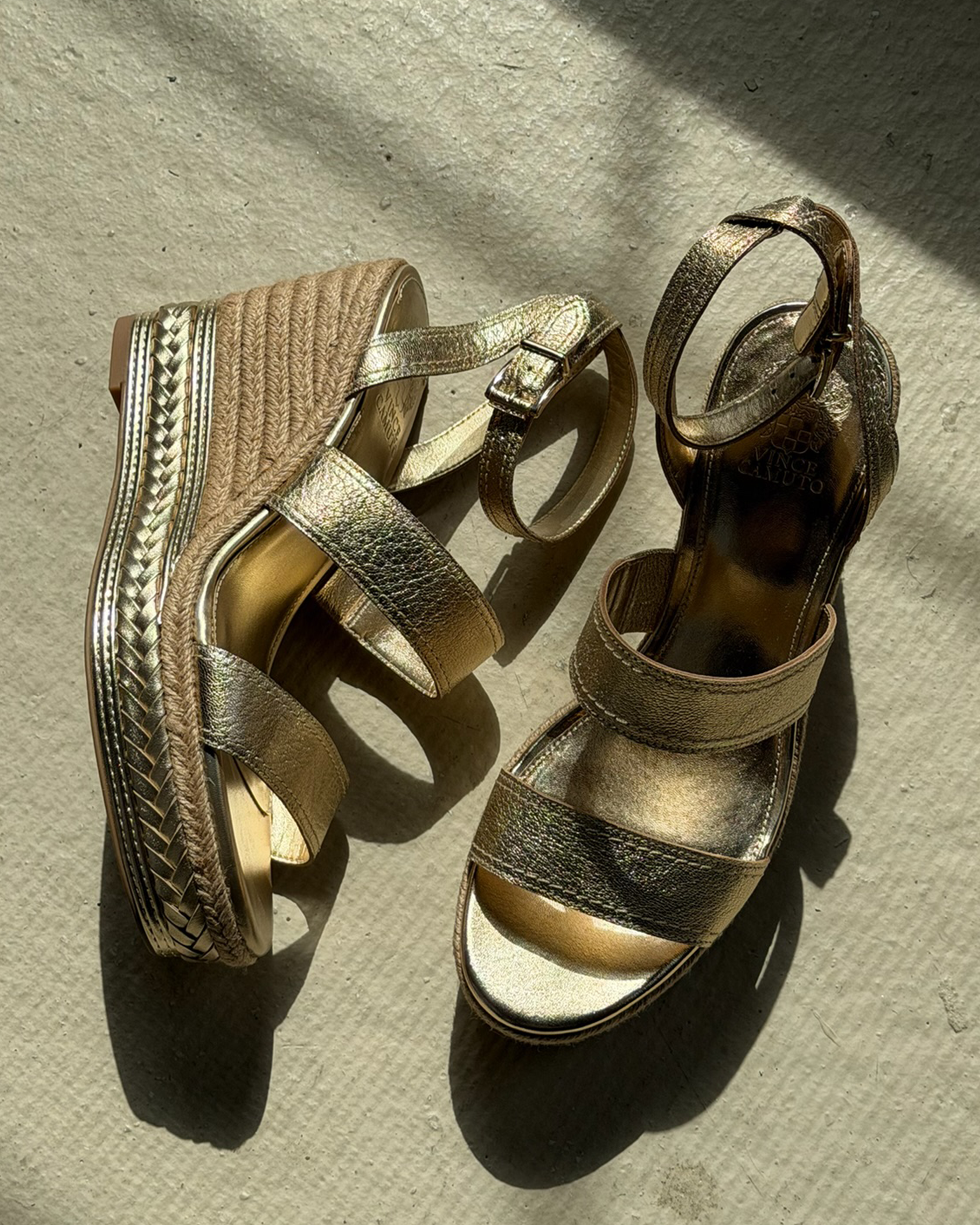 The Vince Camuto Esmia Espadrille Wedge Sandal features gold ankle straps and a braided espadrille platform, shown on a textured beige surface in sunlight.