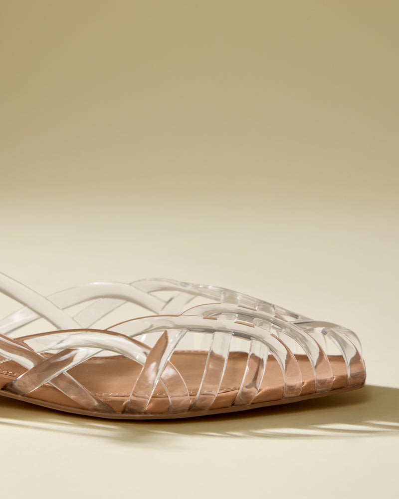 A close-up of the Vince Camuto Stiorra Sandal, featuring transparent crisscross straps, a light brown sole, and a secure slingback strap, displayed on a neutral beige background.