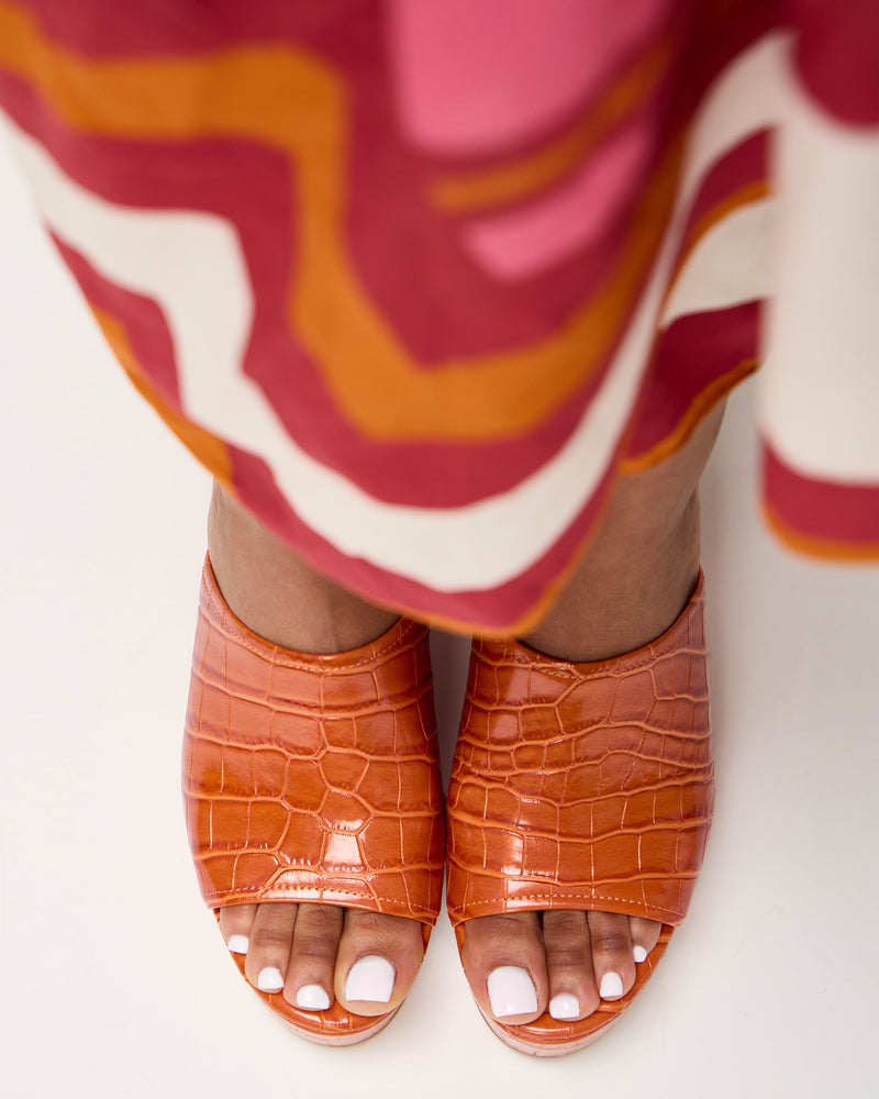 A person stands on a white surface in a colorful patterned skirt and Vince Camuto Etta Wedge Sandals—orange, crocodile-textured leather with open toes and white toenail polish, channeling Y2K style.