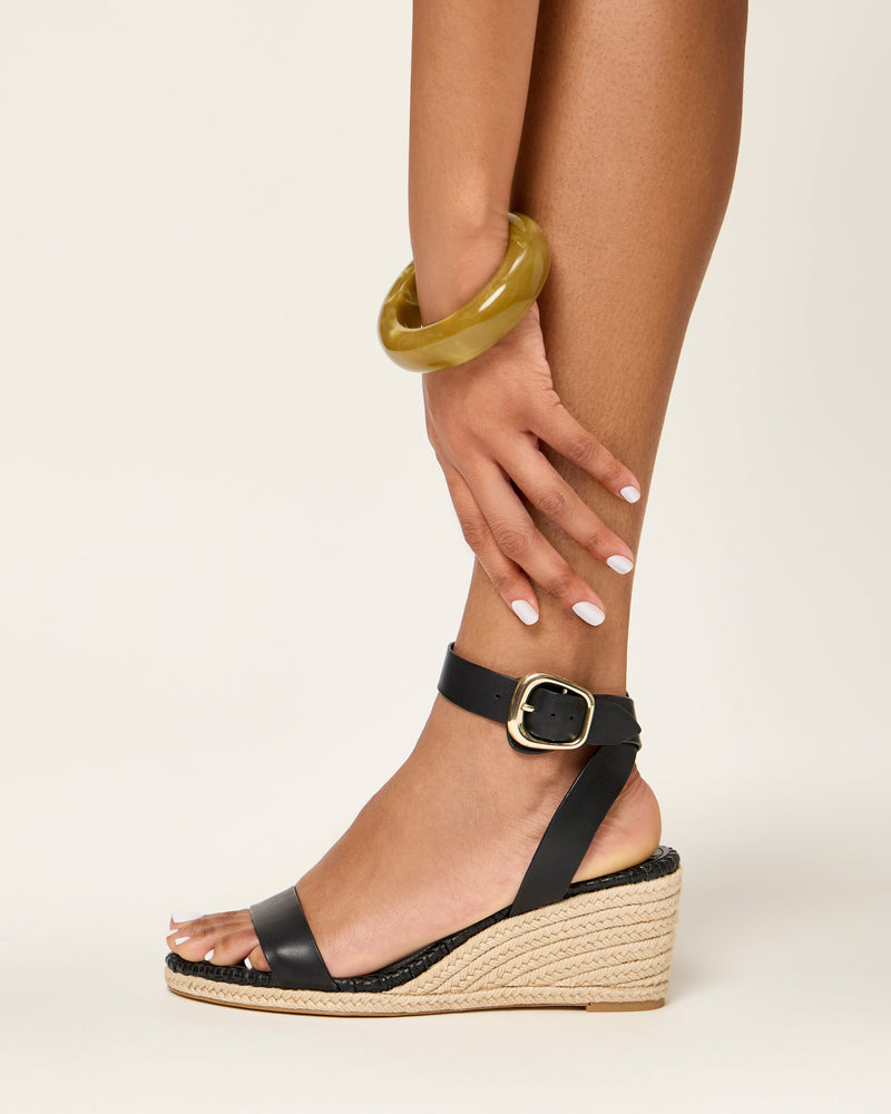A person with light brown skin wears Vince Camuto’s Fainel Espadrille Wedge Sandal, a chunky yellow bangle, and shows off white manicured nails, resting a hand on their shin against a light background for a summery look.