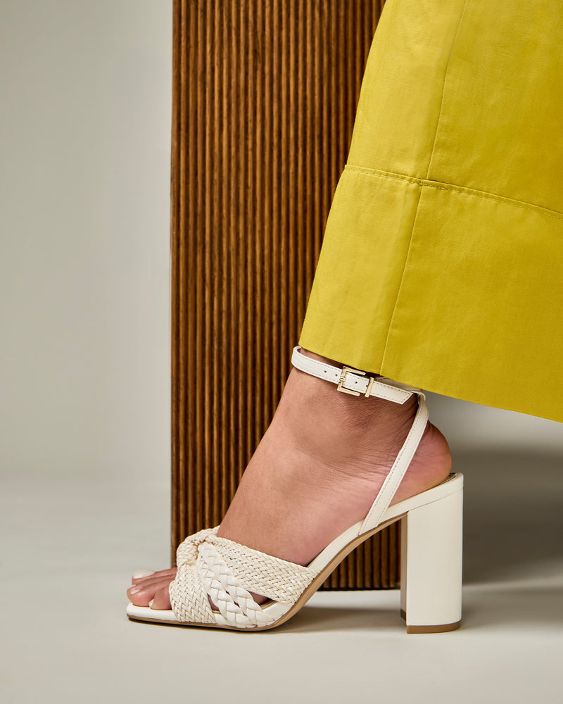 A person in a long yellow skirt wears the Vince Camuto Hanlea Sandal, a white woven high-heeled design with an ankle strap, while standing next to a textured wooden panel.