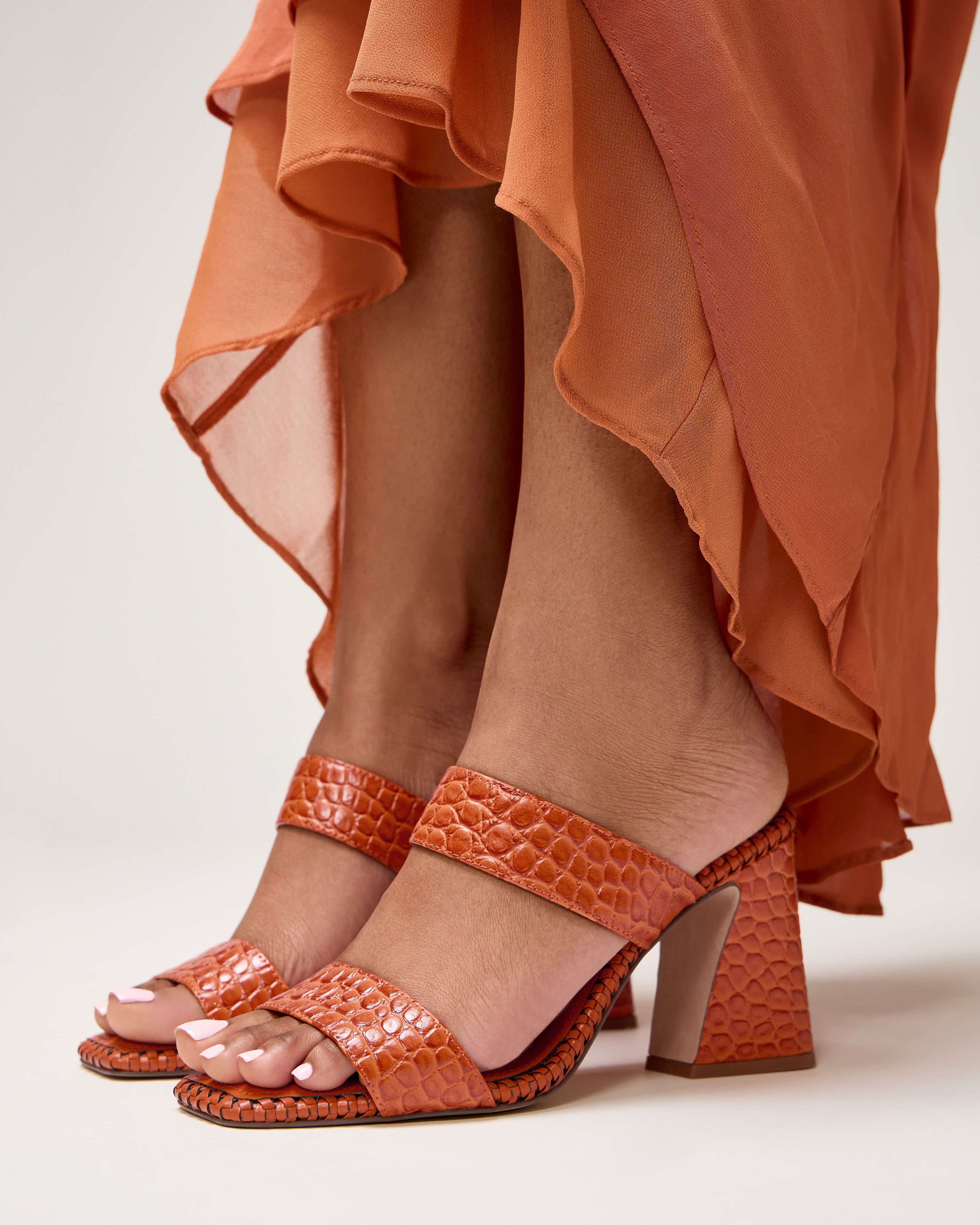 A person stands on a light background wearing an orange, flowy dress and Vince Camuto Kaden Mules—boho-inspired orange textured block heel sandals with a woven midsole.