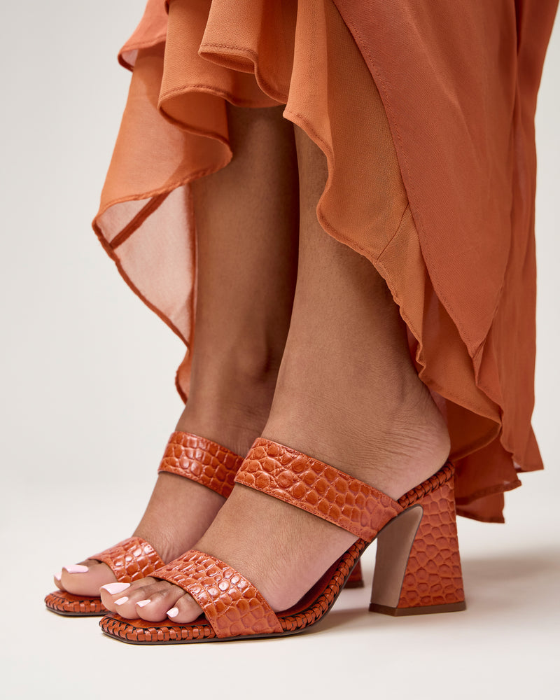 A person stands on a light background wearing an orange, flowy dress and Vince Camuto Kaden Mules—boho-inspired orange textured block heel sandals with a woven midsole.