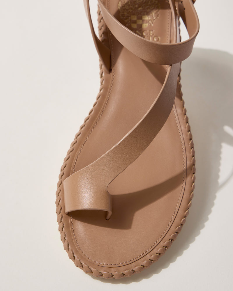 A close-up of the Vince Camuto Libahs Sandal—a tan flat sandal featuring a toe loop, braided edge detailing, wide crisscross leather straps, and an adjustable ankle strap—shown against a light background.