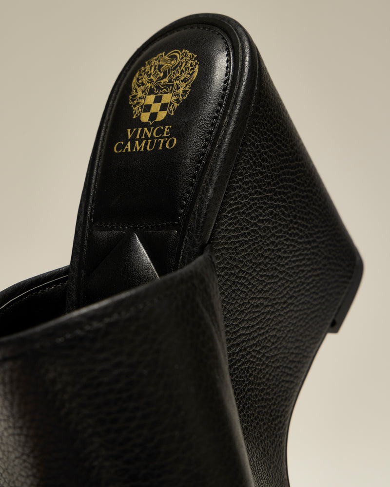A close-up of the black textured Etta Wedge Sandal by Vince Camuto shows the brand’s gold logo and crest on the insole.