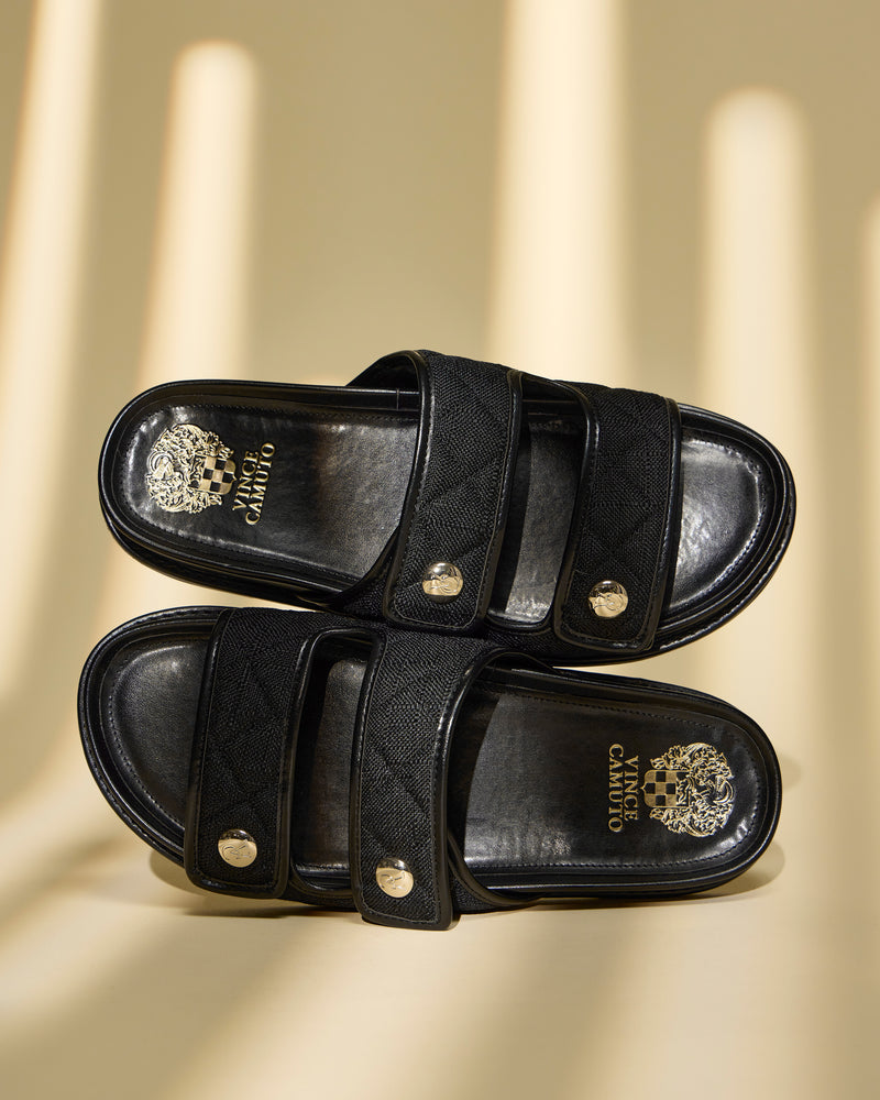 The Vince Camuto Graysin Slide is a black double strap sandal with a quilted upper, gold-toned buttons, and gold brand logos on the insoles, shown on a beige surface with soft striped shadows.