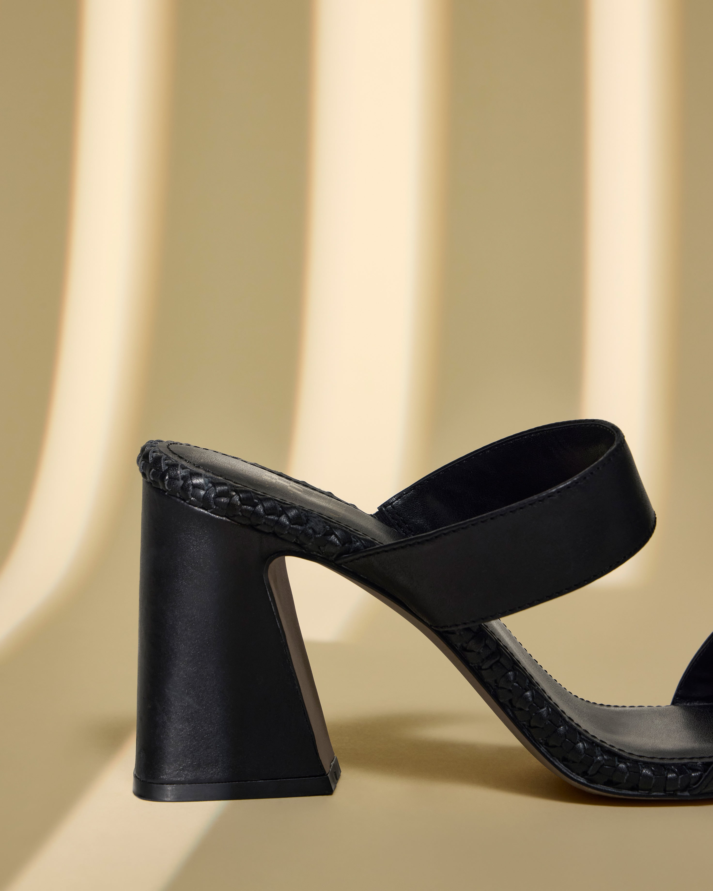 The Vince Camuto Kaden Mule, a black block heel sandal with a woven midsole and wide strap, is shown against a beige background with soft vertical shadows. The photo displays only the rear half of this boho-inspired shoe.