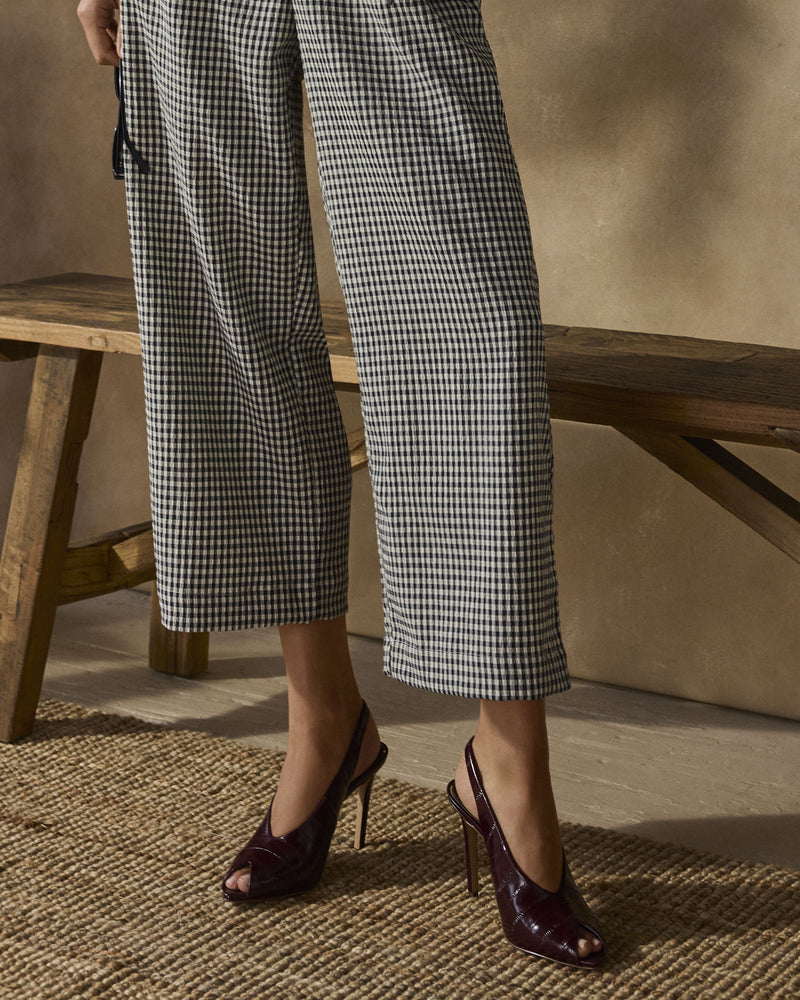 A person wearing black-and-white checkered pants and Vince Camuto Helenza Pumps stands beside a wooden bench on a woven rug.