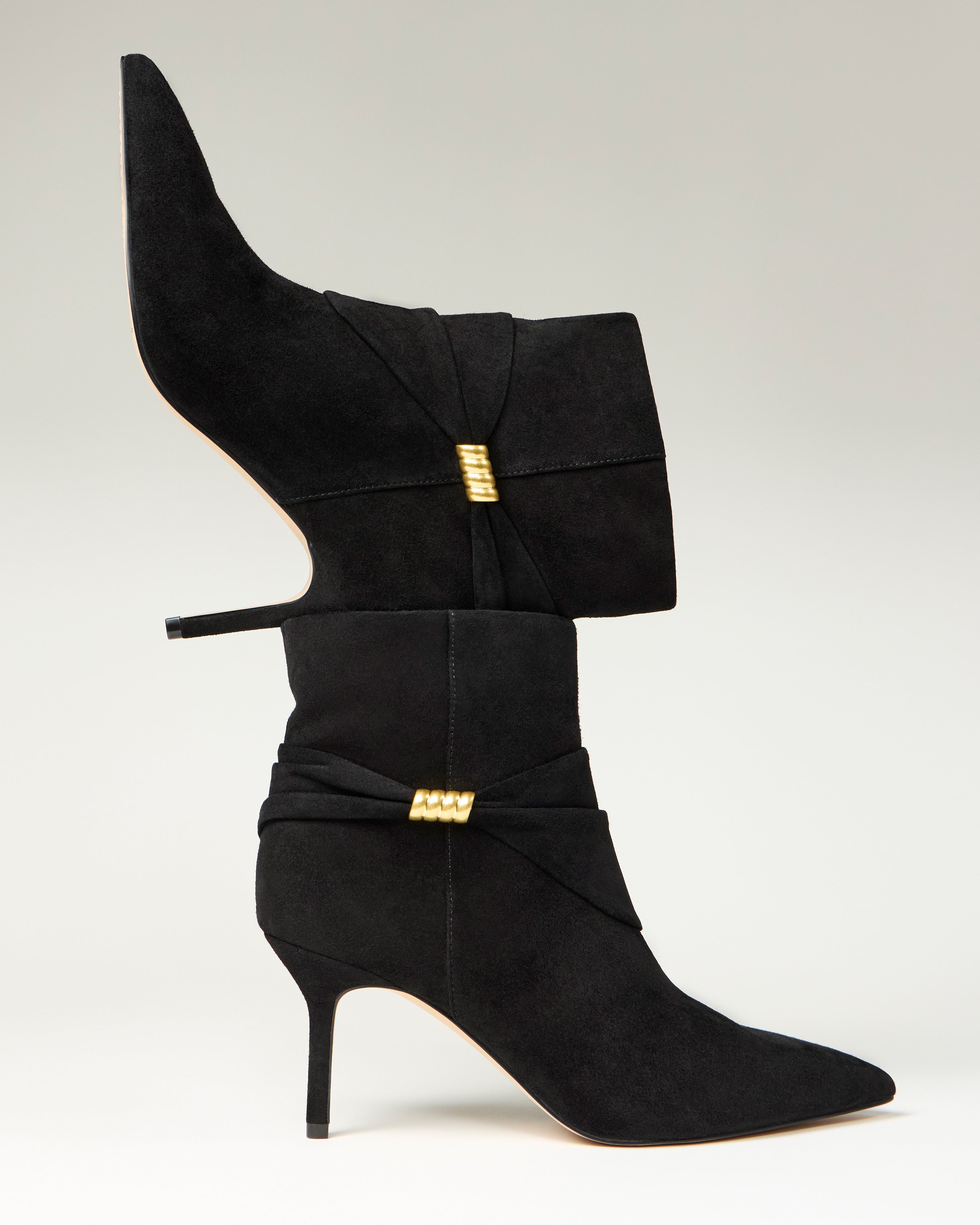 Two Murlea Bootie heels by Vince Camuto, in black suede with gold accents and pointed toes, are stacked vertically—one boot heel-down on the toe of the other—set against a neutral background.