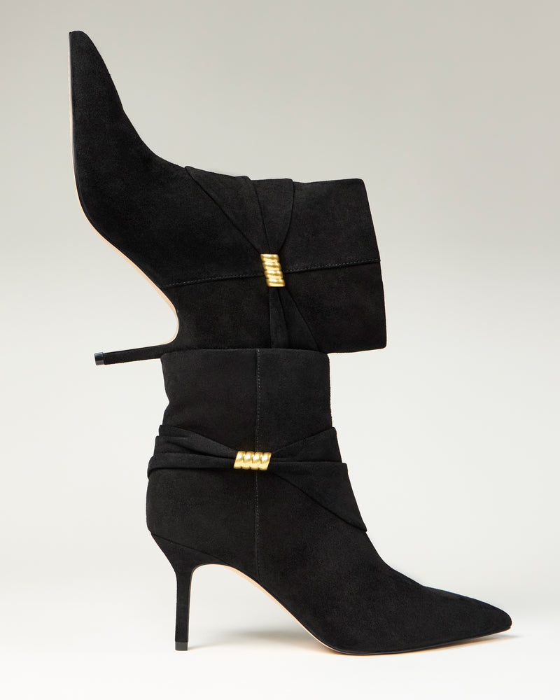 Two Murlea Bootie heels by Vince Camuto, in black suede with gold accents and pointed toes, are stacked vertically—one boot heel-down on the toe of the other—set against a neutral background.