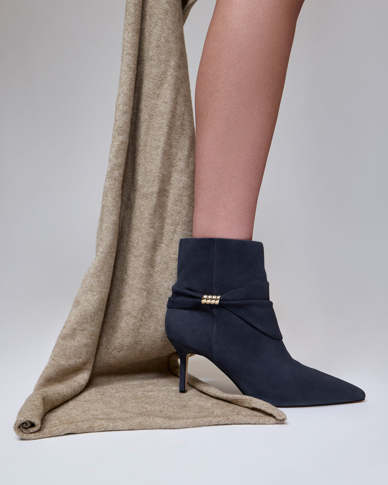 A person in the Vince Camuto Murlea Bootie—navy blue with a gold detail—stands on taupe fabric, with only their lower leg visible against a plain, light background.