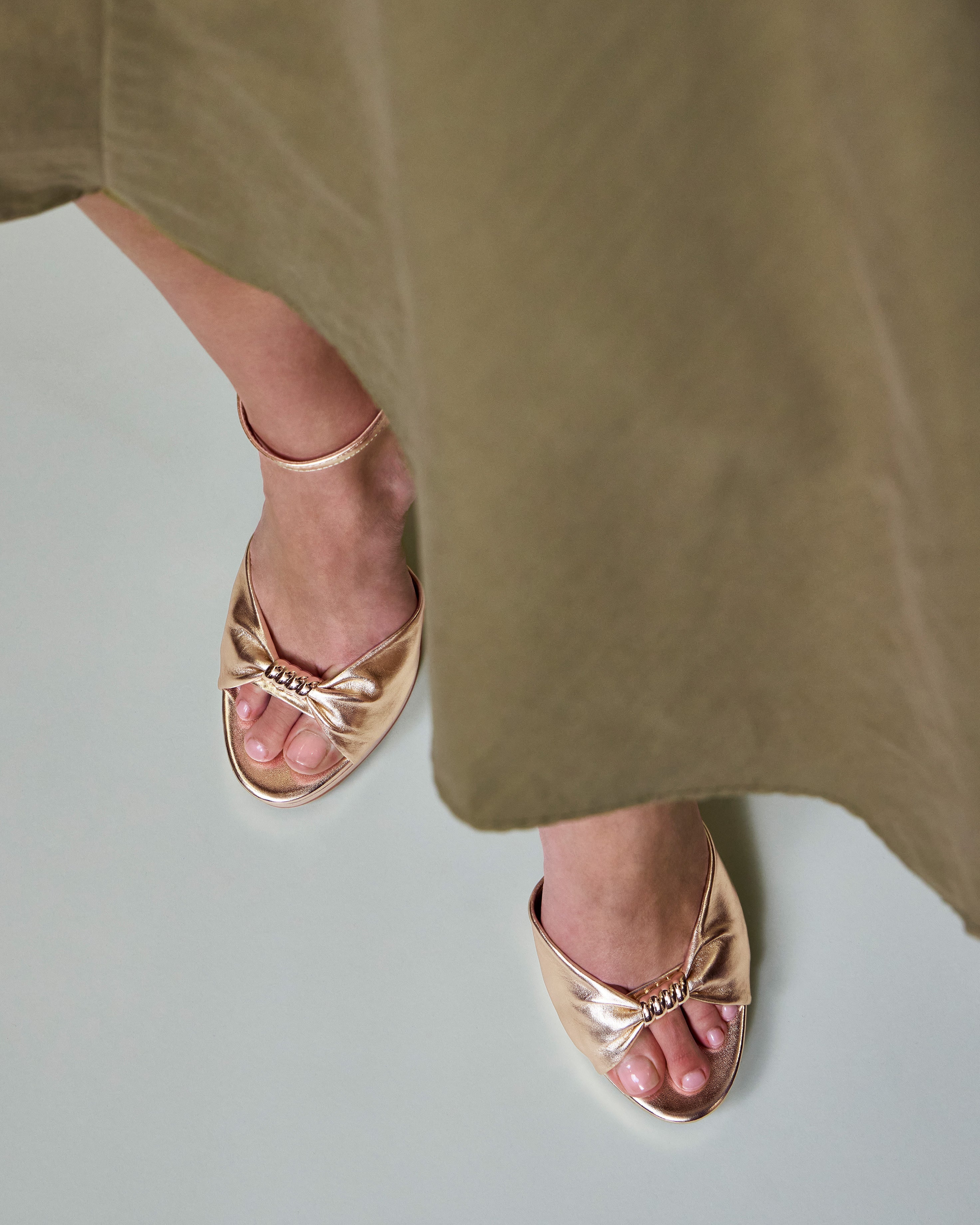 A person in a khaki skirt stands on a light floor wearing Vince Camuto’s Neala Sandal—gold, open-toed, with skinny ankle straps and adorned with bow and jewel details.