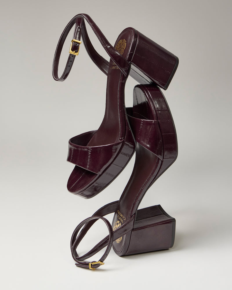 The Vince Camuto Pendreya Platform Sandals, featuring dark burgundy block heels, ankle straps, and gold buckles, are stylishly balanced atop one another against a neutral background.