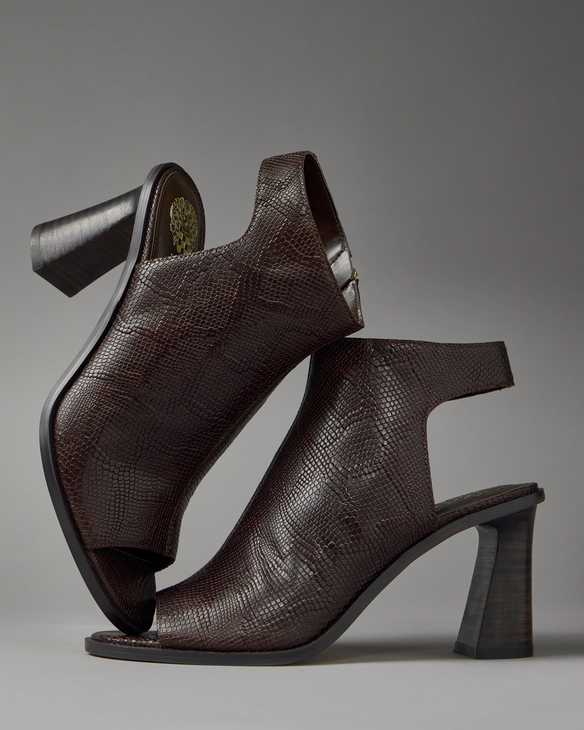 Two dark brown Rimdia Sandals by Vince Camuto, featuring a peep toe, textured reptile-like pattern, and block heels. One stands upright while the other is balanced on its heel to show the sole and gold emblem against a plain gray background.