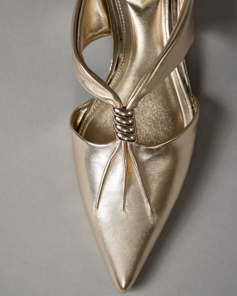 Close-up of the Vanika Mule by Vince Camuto, featuring a metallic gold pointed toe, pleated details, and a twisted strap design over the vamp, displayed against a neutral gray background.