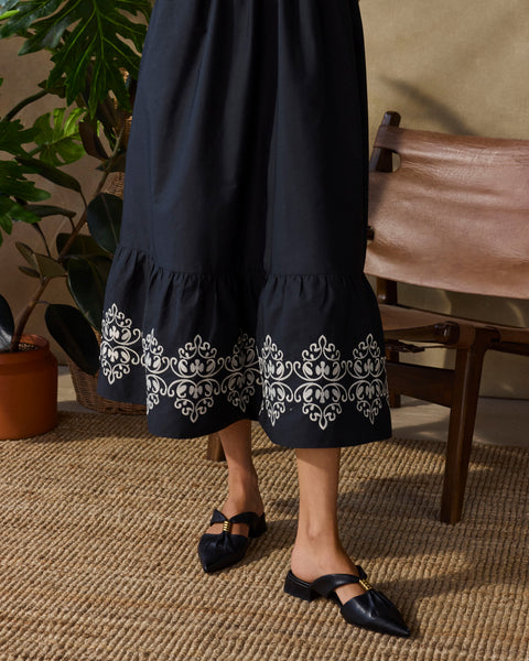 A person stands on a woven rug wearing a long black skirt with white embroidery near the hem and black Vanika Mules by Vince Camuto; a wooden chair and green potted plants are in the background.