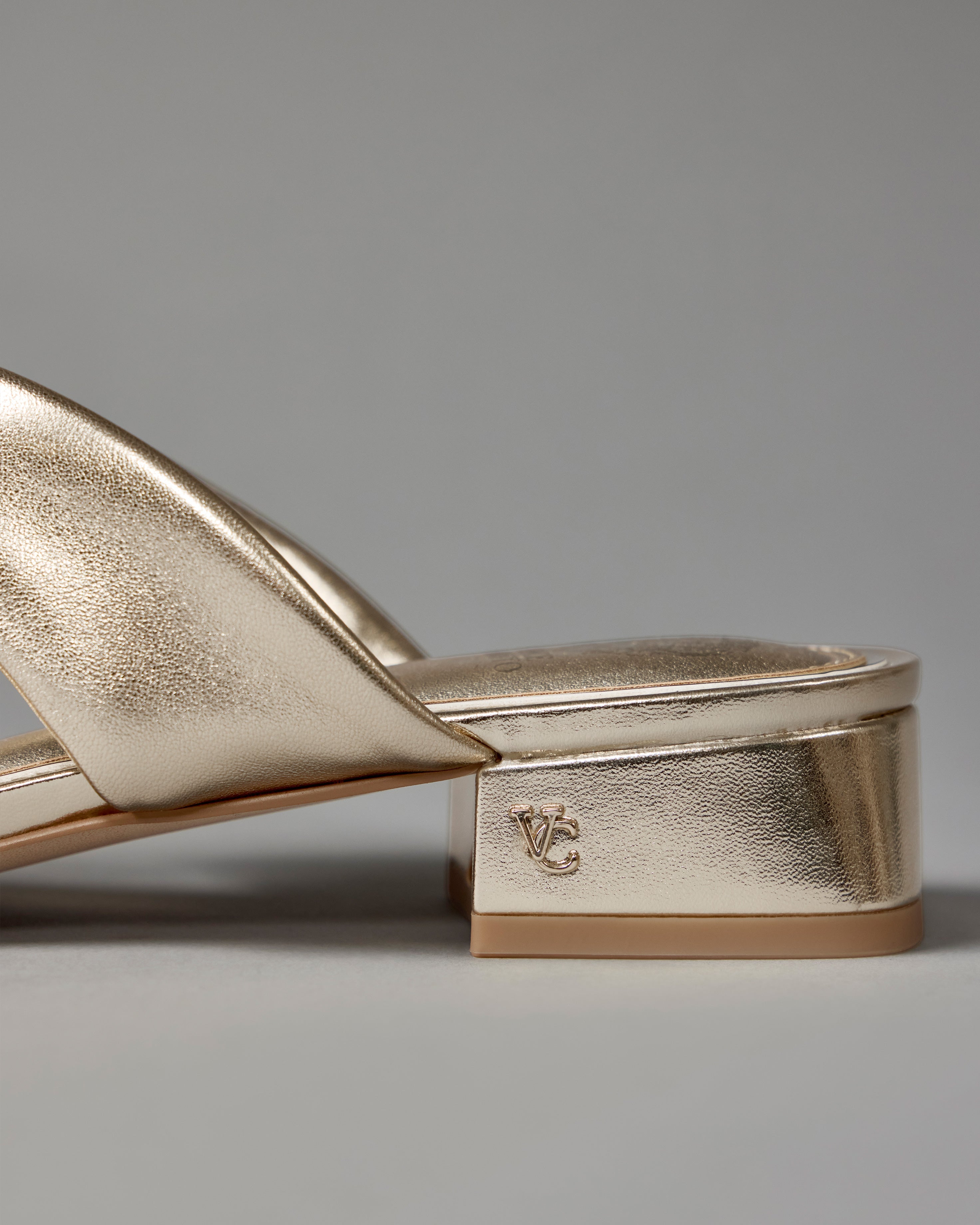 Close-up of the Vince Camuto Vanika Mule in gold metallic with a low block heel featuring the VC logo, set against a plain gray background.