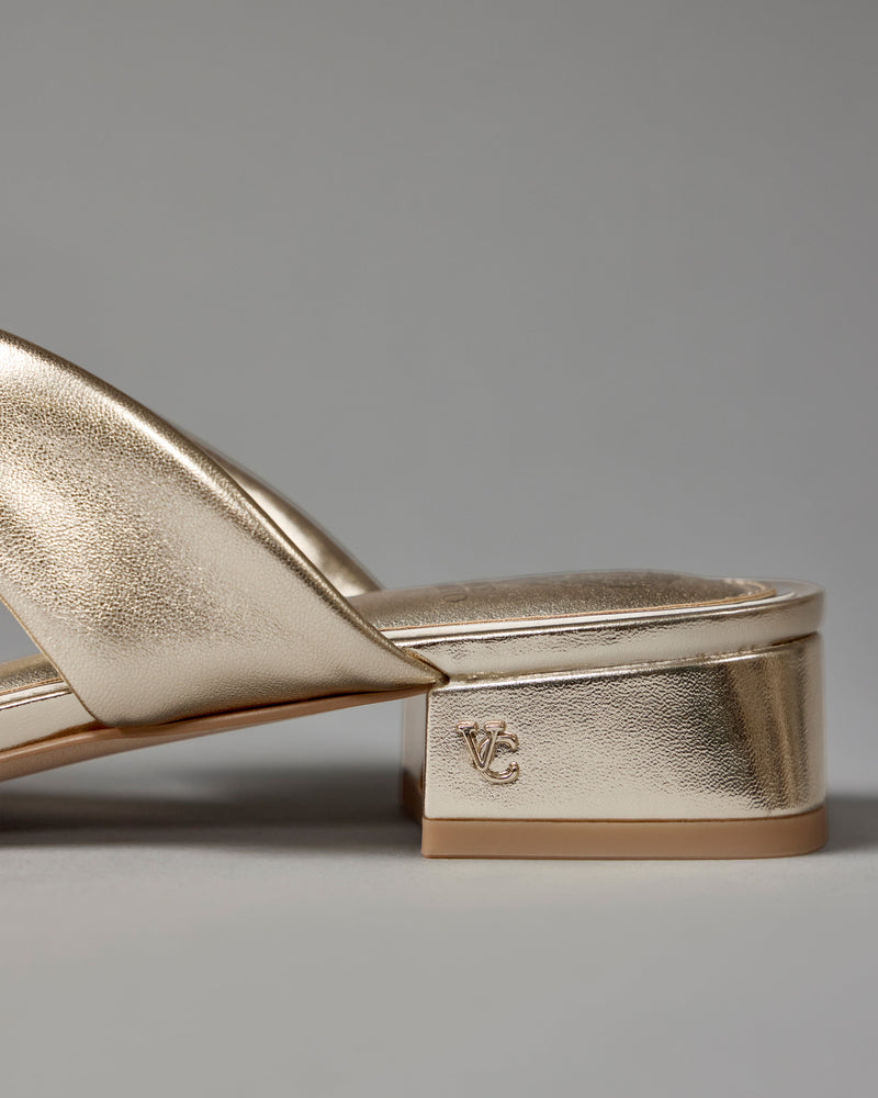 Close-up of the Vince Camuto Vanika Mule in gold metallic with a low block heel featuring the VC logo, set against a plain gray background.