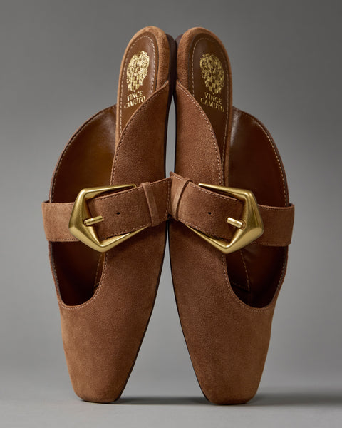 The Vellma Buckle Mule by Vince Camuto features brown suede, pointed toes, gold buckles, and embossed gold branding on the insoles, shown upright against a neutral gray background.